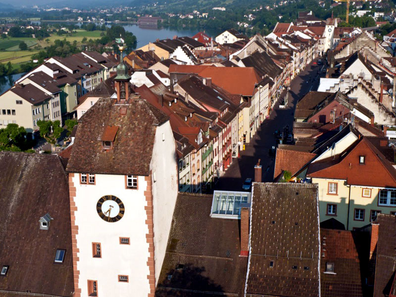 Glockenturm mit Uhr. Im Hintergrund die Stadt Waldshut mit mehreren Häusern.